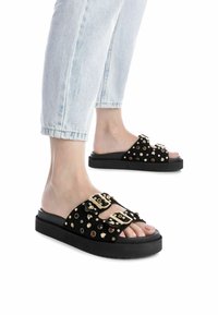 Sandali neri con parte superiore in suede, caratterizzati da borchie e fibbie in tonalità oro, su una spessa suola piatta nera, abbinati a jeans chiari.