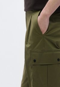 Olivzaļas krāsas darba bikses ar sānu kabatu, kas aiztaisāma ar atloku un melnu pogu. Izgatavotas no gludas, izturīgas auduma ar pieguļošu piegriezumu.