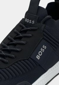 Navy sneaker featuring a textured mesh upper, intricate lacing system, rubber sole, and contrasting white accents with "BOSS" logo detail.