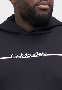 Zwarte hoodie van een katoenmix. Voorzien van een witte "Calvin Klein" logo met daaronder een horizontale streep, wat een stevige textuur laat zien.