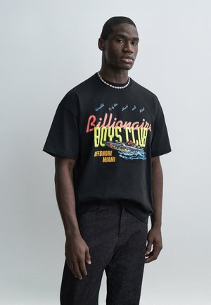 Hombre que lleva una camiseta negra de gran tamaño con un gráfico colorido que dice "Billionaire Boys Club Offshore Miami" y pantalones vaqueros negros, posando frente a un fondo gris.