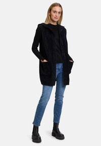 Schwarze, strukturierte Weste mit Kapuze, ausgestattet mit einer Knopfleiste und zwei Seitentaschen, getragen über einem schwarzen Oberteil und blauen Jeans. Schwarze, klobige Stiefel.