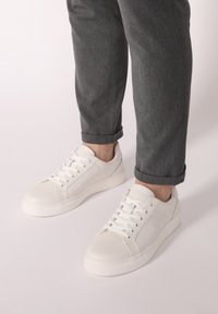 Weiße Leder-Sneaker mit einer glatten Oberfläche, strukturierten Seitenpaneelen und einer dicken Gummisohle. Die Schnürsenkel verlaufen durch sechs Ösen für einen sicheren Sitz.