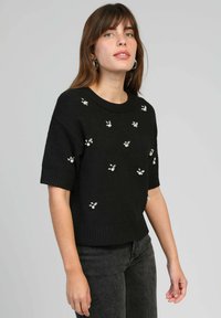 Pull en maille noir à manches courtes, orné de perles en formes florales. Encolure et ourlet côtelés. Assorti à un jean foncé.