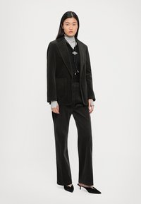 Dark green corduroy suit with a fitted blazer, wide-leg trousers, and silver hardware accents. Layered over a gray turtleneck.