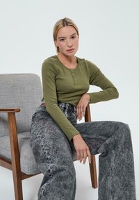 Top à manches longues vert olive avec des détails de boutons, associé à un jean en denim gris taille haute et à coupe ample. Le tout devant une chaise grise simple.