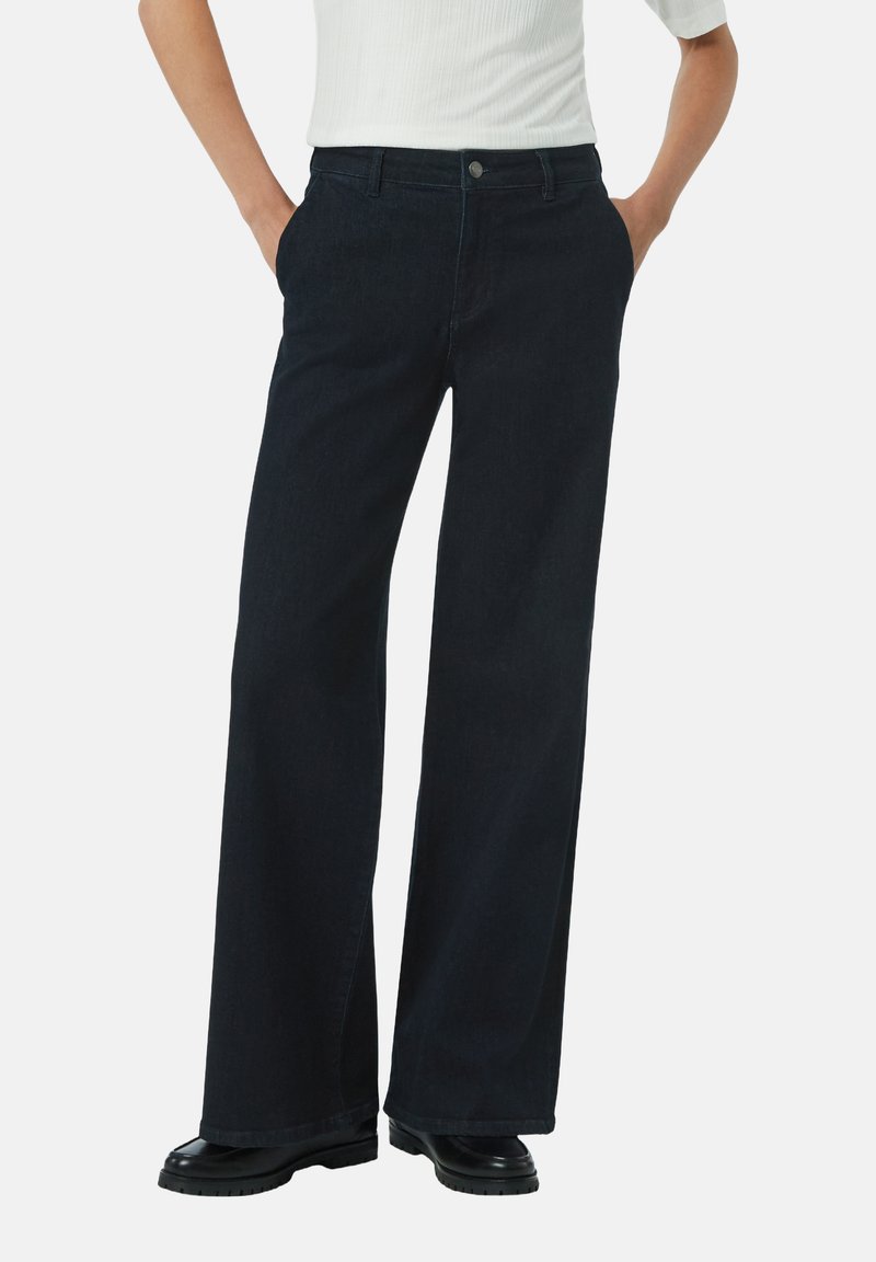 Szerokie ciemnoniebieskie jeansy z wysokim stanem, z zapięciem na guzik i bocznymi kieszeniami, w połączeniu z czarnym obuwiem.