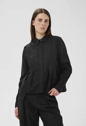 Kobieta o prostych brązowych włosach, ubrana w czarną, zapinaną na guziki bluzkę z długim rękawem oraz pasujące czarne spodnie na jasnym, jednolitym tle.