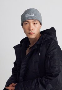 Cappello di lana grigio con etichetta, indossato con un giubbotto imbottito nero con fodera in shearling e cordino. Outfit casual e a strati.