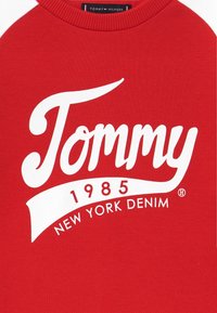 Rotes Sweatshirt mit weißem Aufdruck: „Tommy 1985 New York Denim.“ Verfügt über einen gerippten Rundhalsausschnitt und kontrastierende weiße Ärmel.