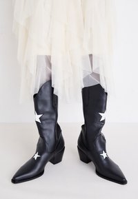 Czarne skórzane kowbojki z białymi gwiazdkami, spiczastymi noskami i średnią blokową piętą, na tle jasnej warstwowej tiulu.