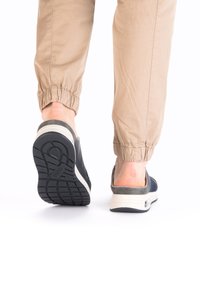 Des chaussures noires sans lacets avec un dessus texturé et un talon gris contrastant. Elles possèdent une semelle blanche rembourrée et une semelle à motif. Pantalons beiges à revers.