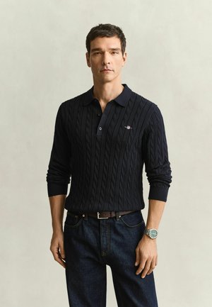 Mann trägt ein schwarzes, kabelgestricktes, langärmeliges Poloshirt, dunkle Jeans, einen braunen Gürtel und eine Armbanduhr und steht vor einem schlichten, hellen Hintergrund.