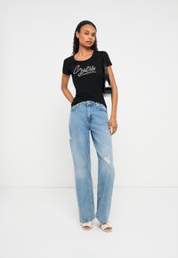 Kobieta ubrana w czarną koszulkę "Guess", jasnoniebieskie, poszarpane dżinsy w szerokim kroju, białe sandały na obcasie z odkrytymi palcami oraz czarną torebkę na ramię, stojąca przy białej ścianie.