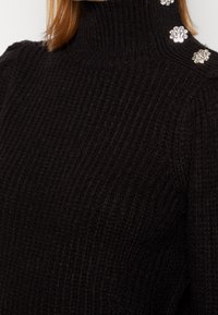Maglione nero a coste con colletto a dolcevita, spalle imbottite e tre bottoni decorativi a forma di fiore in strass sulla spalla sinistra.