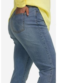 Jeans de mezclilla azul con forma ajustada, que incluyen un bolsillo trasero y detalles de costura sutiles. Llevados con una camisa amarilla.