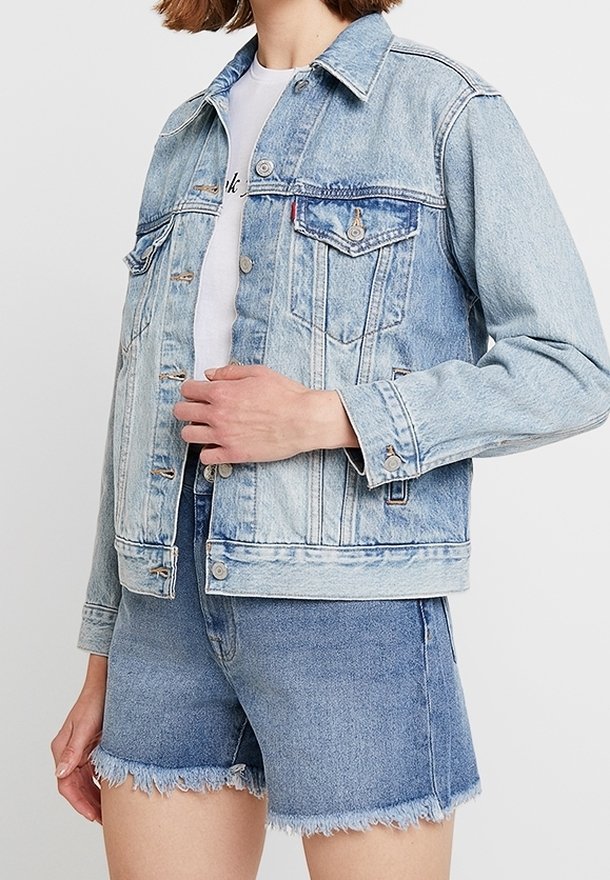 Personne portant une veste en jean bleu clair et un short en jean effiloché, avec une chemise blanche partiellement visible sous la veste.