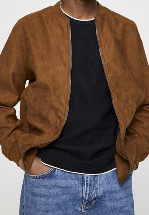 Personne portant une veste bomber en daim marron par-dessus un pull en maille noir et un jean bleu, les mains dans les poches de la veste, fond neutre.