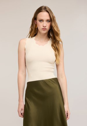 Ung kvinde med langt, bølget hår iført en ærmeløs hvid top med bølgede kanter og et olivengrønt satin-nederdel, stående mod en neutral baggrund.