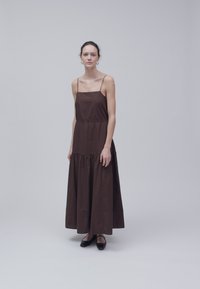 Femme debout dans une robe longue marron foncé sans manches avec de fines bretelles et des ballerines noires, sur un fond uni gris clair.