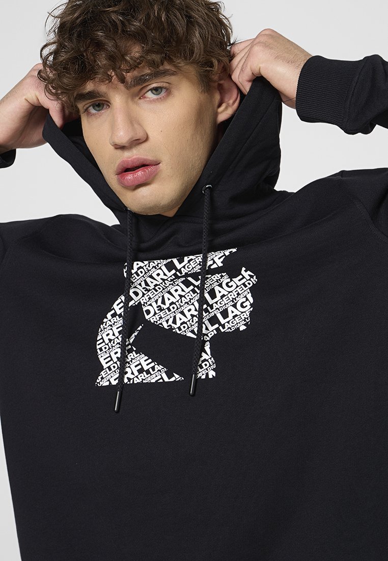 Sweatshirt à capuche noir avec une finition texturée. Présente un grand logo graphique blanc sur la poitrine avec des éléments de texte répétés. Cordons de serrage ajustables.