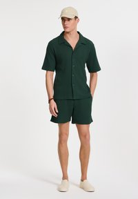 Tummanvihreä ribattua lyhythihaista paitaa nappiedessä, yhdistettynä samanvärisiin shortseihin. Mallilla on päällään vaalea beige lippalakki ja kermaiset liukulumpparit.