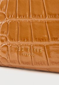 Leren tas in cognackleur met krokodillenprint en een glanzende afwerking, met onderaan het goudkleurige, aangebrachte "Ted Baker"-logo, dat de textuur en het patroon benadrukt.