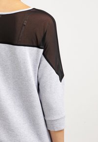 Grijze sweatshirt met zwart doorschijnend mesh aan de bovenrug, met driekwart mouwen en een ronde hals. De stof heeft een gladde textuur.