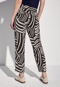 Un pantalon noir et beige à motif présente un design géométrique, une taille haute, une coupe ample et des poches latérales. Un modèle est montré de dos.
