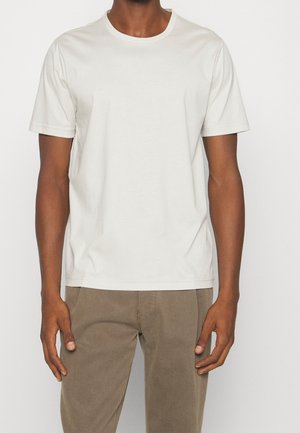 Mann trägt ein einfaches weißes T-Shirt mit kurzen Ärmeln und Rundhalsausschnitt sowie hellbraune Hose, stehend vor einem einfarbigen Hintergrund.