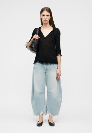 Naine kannab musta V-kaelusega topi, millel on narmad, laia valgelt sinist kulunud teksapükse, musti madalaid kingi ja kannab mustast punutud õlakotti.