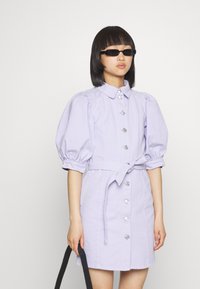 Vestido lilás claro com botões, mangas bufantes, cinto às riscas e botões prateados. Modelo segurando uma bolsa preta, usando óculos escuros escuros.