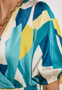 Blusa a maniche corte in tessuto setoso con un motivo geometrico in tonalità di blu, giallo e crema, con vita aderente e maniche arricciate.