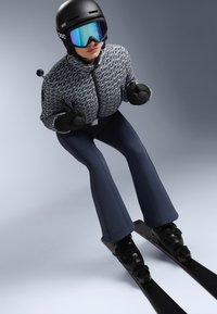 Skijaš u pletenoj, plavoj jakni i crnim skijaškim hlačama, nosi crnu kacigu i zrcalne naočale, na crnim skijama protiv sivo pozadine.