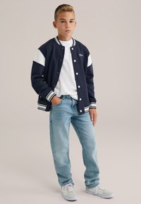 Mörkblå varsityjacka med vita axelpaneler, ribbade muddar och tryckknappar, i kombination med ljusblå jeans och ljusgröna sneakers.