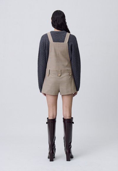 Pantalones de tirantes a cuadros en color beige con tirantes ajustables, combinados con un suéter de punto oscuro y botas de cuero marrón hasta la rodilla. Con frente plano y detalles de botones.