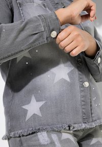 Grå denimjacka med stjärnmönster, silverfärgad knäppning och fransad fåll. Dekorerad med små rhinestones för extra detalj.