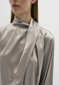 Beige satijnen blouse met een hoge hals, voorzien van gedrapeerde stoffen details en een gladde textuur, die de zachte glans en het elegante ontwerp benadrukt.
