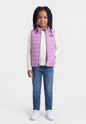 Noor tütarlaps põimitud juustega, kellel on seljas helelilla sulejope, valge pika varrukaga särk, sinised teksad ja valged tennised, seismas.