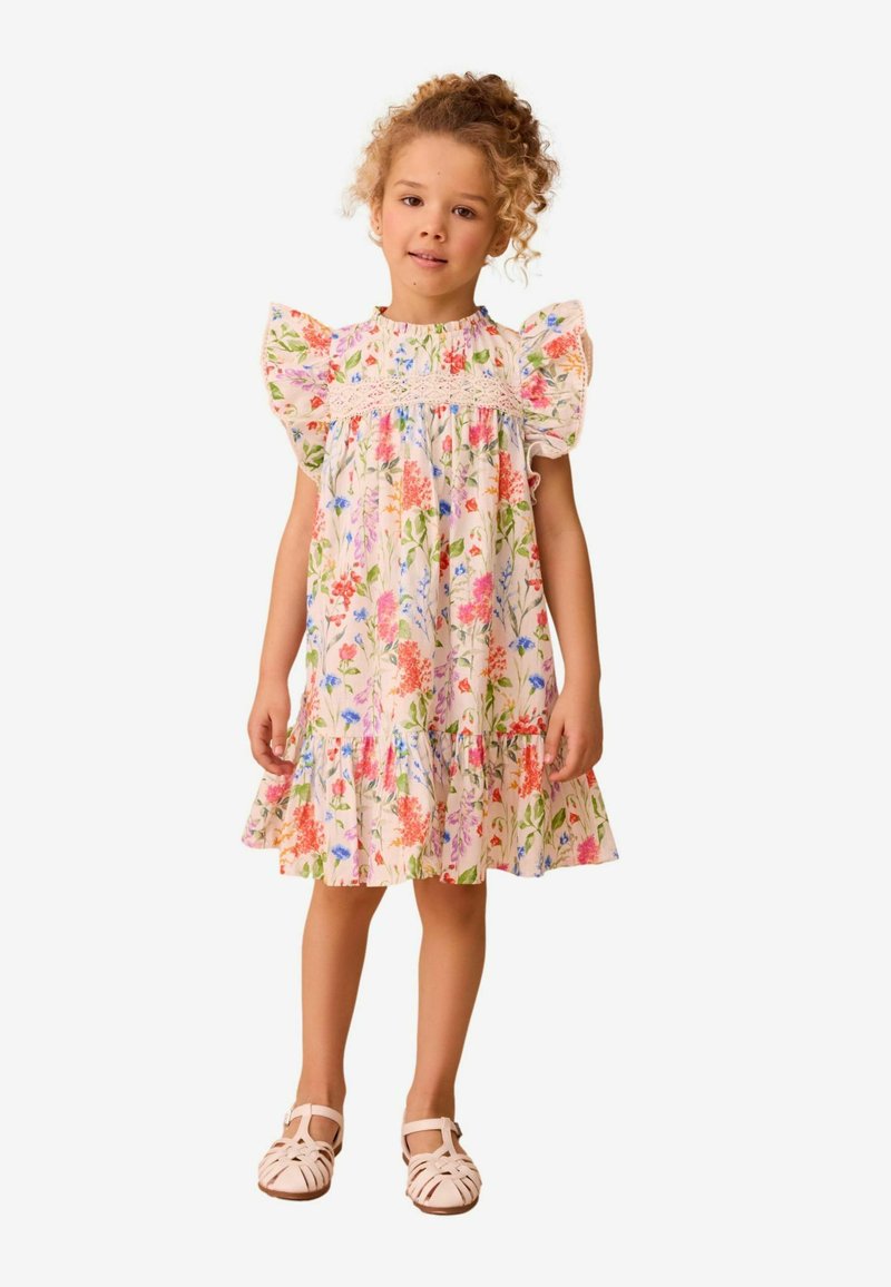 Bloemenjurk met kapmouwen, een gerimpelde zoom en een smock-borstvoeding. Heeft kleurrijke bloemen op een lichte achtergrond. Gecombineerd met zachte, enkelband sandalen.