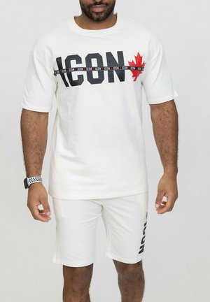 Homme portant un short blanc et un T-shirt blanc surdimensionné assorti avec le texte noir audacieux "ICON" et une feuille d'érable rouge sur la poitrine et le short.