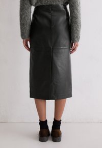 Czarna skórzana spódnica midi o gładkiej fakturze, z tylnym rozcięciem i ukrytym suwakiem, zestawiona z ciemnymi skarpetkami i brązowymi butami.