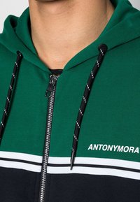 Felpa con cappuccio verde e blu navy, dotata di zip frontale, lacci neri con dettagli bianchi e un logo bianco sul petto.