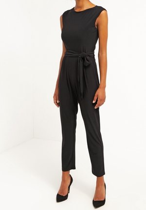 Kvinde iført en sort ærmeløs jumpsuit med et bundet bælte i taljen og sorte spidse høje hæle, stående mod en ensfarvet hvid baggrund.