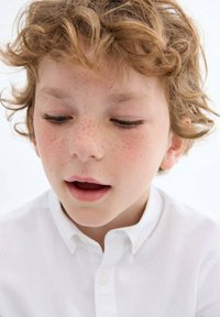 Garçon avec des cheveux blonds bouclés et des taches de rousseur portant une chemise blanche à boutons. Il regarde vers le bas, avec une expression neutre sur son visage.