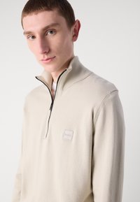 Sweater bege com fecho zip e gola canelada, e um pequeno logótipo no peito. Textura suave e ajuste descontraído, projetado para uso casual.