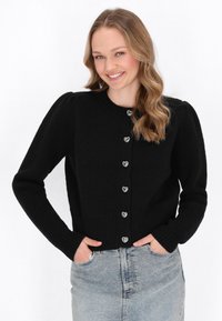 Černý pletený cardigan s vroubkovaným lemem, dlouhými rukávy a stříbrnými knoflíky ve tvaru srdce vpředu. Modelka ho nosí přes světle modrou džínovou sukni.