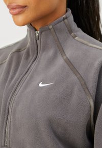 Szara polarowa kurtka z wysokim kołnierzem, zamek błyskawiczny z przodu oraz beżowymi akcentami wzdłuż szwów; zawiera małe białe logo Nike.