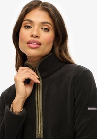 Veste en polaire noire avec un col haut, dotée d'une fermeture éclair dorée et d'accents vert olive. Patch logo sur la manche gauche. Texture lisse.