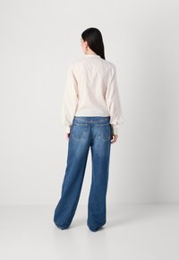 Una persona con lunghi capelli scuri è di spalle, indossando una blusa leggera e svolazzante e jeans blu larghi, in piedi contro uno sfondo grigio semplice.